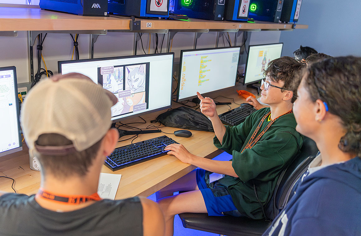 A group of people sitting at computer workstations, one pointing at a screen displaying a comic strip while others look on.