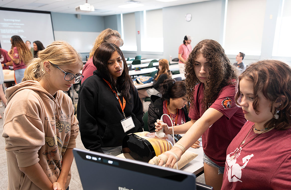 A group of students in a classroom setting gathered around a table with medical equipment, including an ultrasound machine.