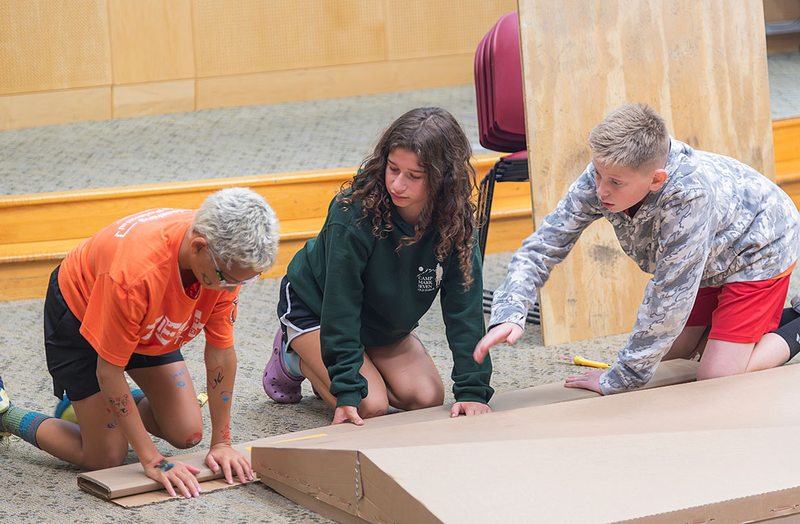 Three students kneeling on the floor, collaborating on a large piece of cardboard during a group activity. They are dressed in casual clothing including an orange shirt, green sweatshirt, and camouflage hoodie.