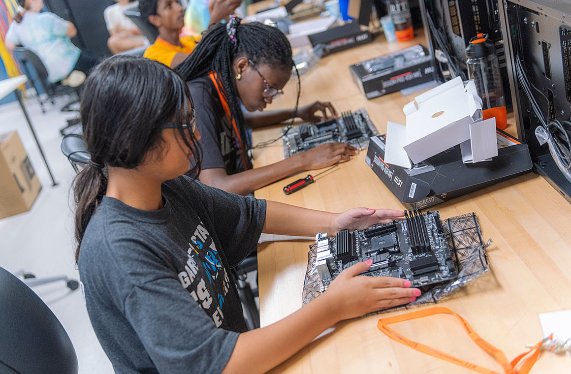 Individuals seated at a workbench assembling computer motherboards with tools and components, including boxes and screwdrivers, in a technology lab setting.