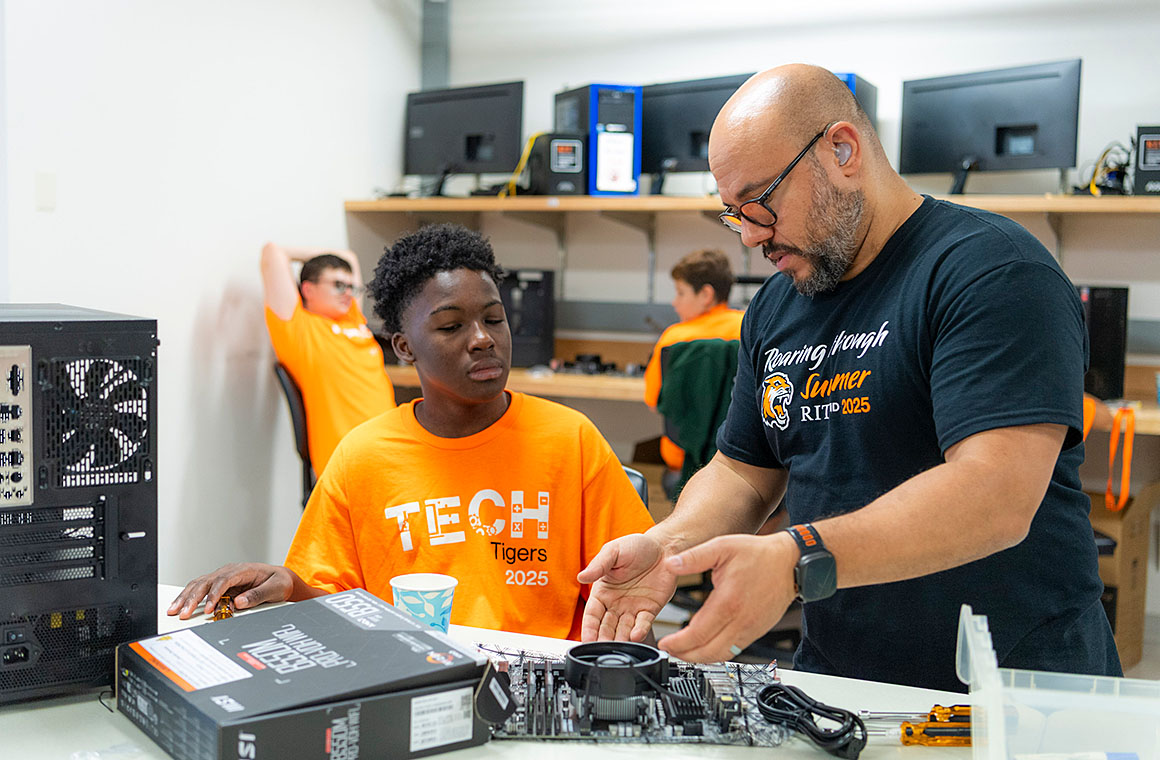 A teacher instructing a student on computer hardware assembly in a classroom with other students working on computers in the background.