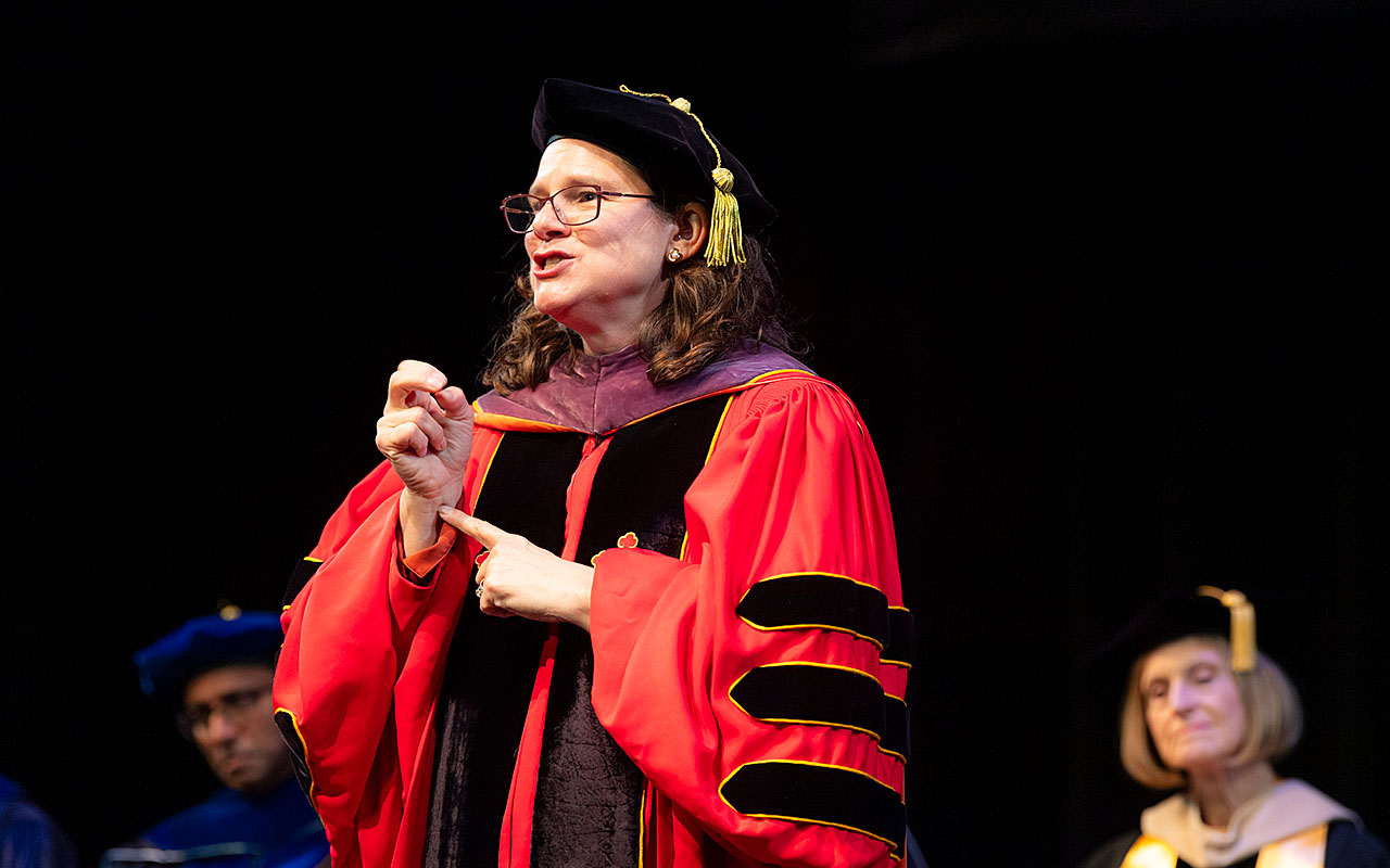A person in academic regalia, wearing a red gown with black and gold accents and a black cap with gold tassels, speaking on stage with others in similar attire in the background.