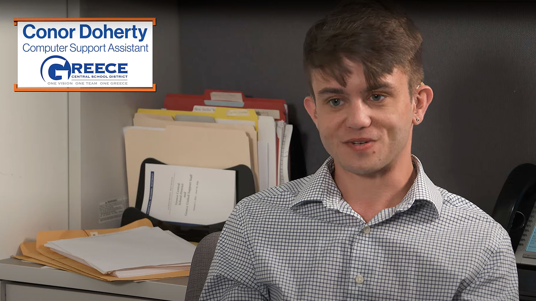 Conor Doherty, Computer Support Assistant at Greece Central School District, sitting in an office with folders and documents behind, wearing a checkered shirt and speaking.