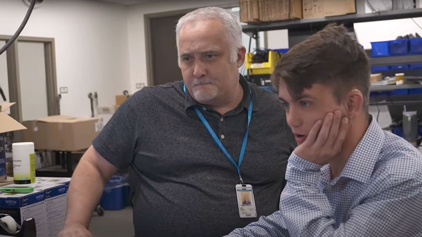 Two people looking at a computer screen in an office or workshop; one with gray hair and a lanyard, the other with short brown hair resting their hand on their face.