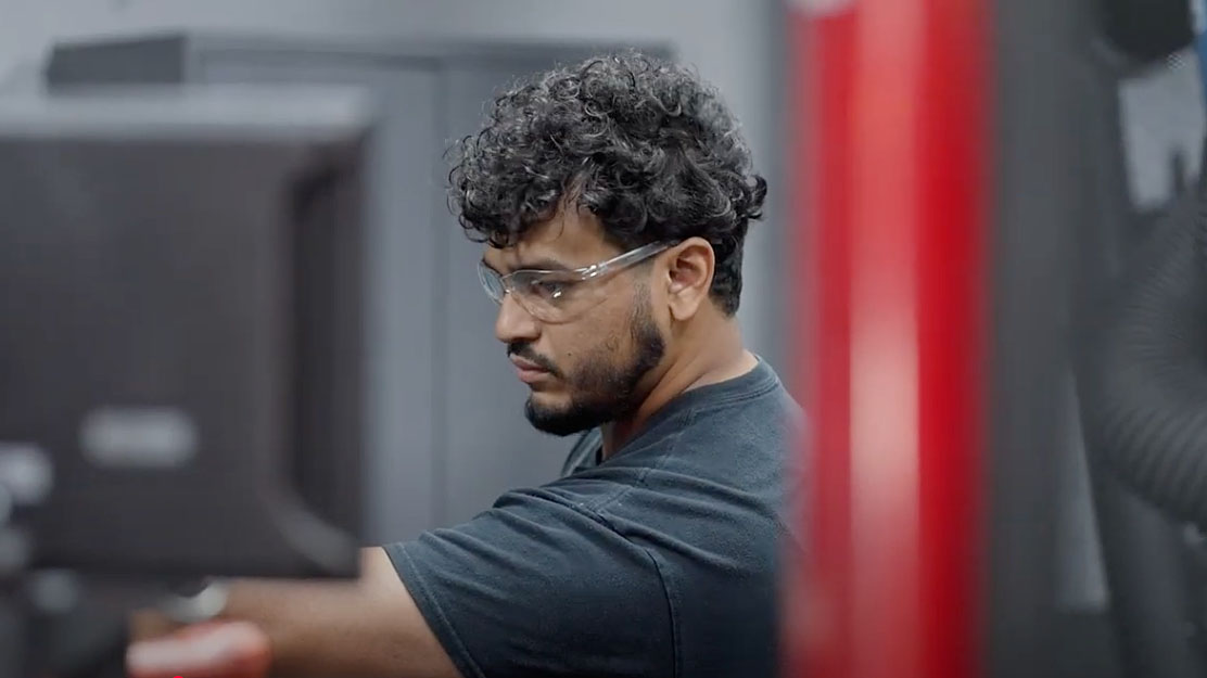 Person with curly hair and safety glasses working in a technical or industrial environment, focused on a task.