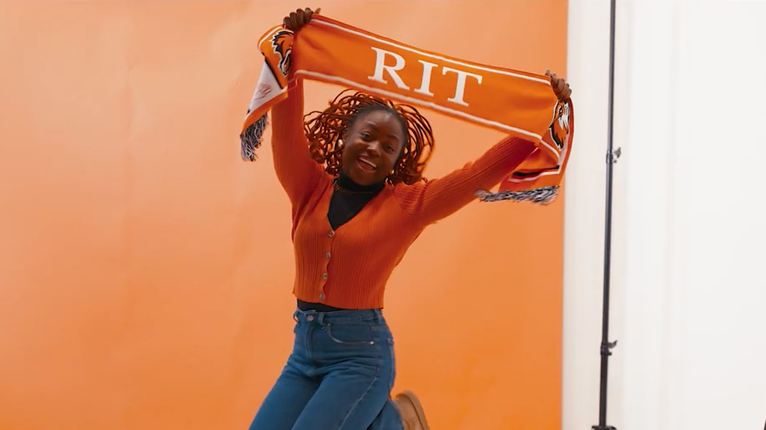 A young Black woman with long braids jumps joyfully in front of an orange background, holding an orange and white scarf above her head that reads RIT. She is wearing an orange ribbed cardigan and blue jeans.