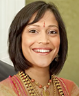 Sasha, a brown Indian woman, has brown eyes and medium length dark brown hair with bangs. She is wearing rose-gold saree with traditional Indian necklaces and a bindi. Sasha is smiling in front of a blurred olive green and white backdrop.