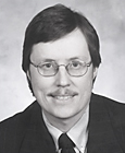 Photo of man with dark hair, glasses, wearing dark suit and tie and light shirt