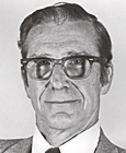 Photo of man with dark hair and glasses, wearing dark suit, white shirt, and tie.