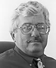 Photo of man with white hair, glasses, wearing dark shirt and neck tie.