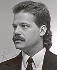 Photo of man 3/4 view, with dark hair and mustache, wearing suit and tie.