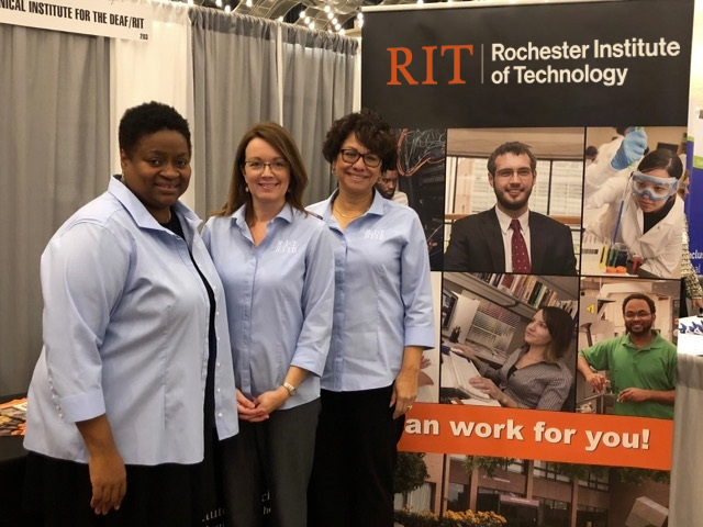 Three women standing next to NCE display at a conference