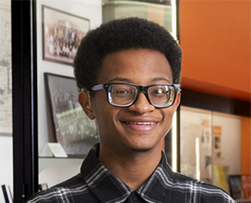 Young man, smiling, dark hair, glasses, wearing check shirt and white pants, hands crossed over waist.