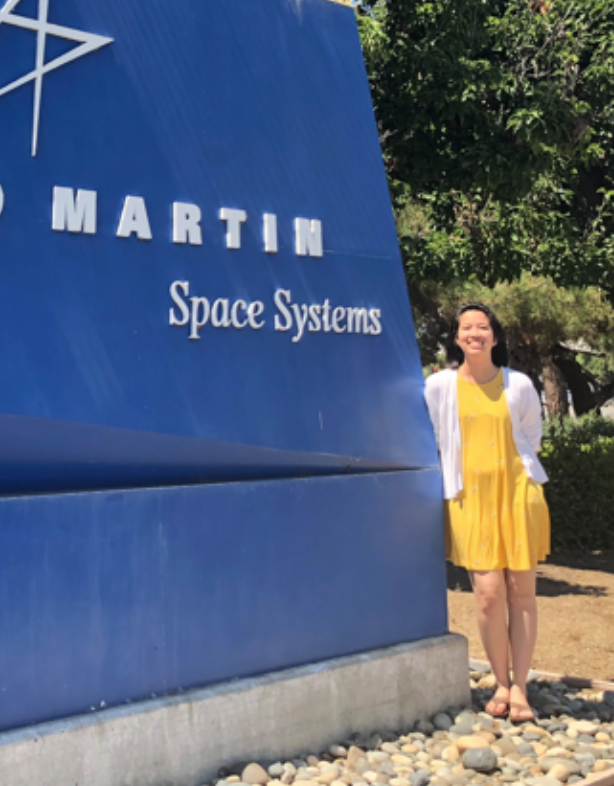 Photo of Amanda Bui Photo of Amanda Bui, young woman with dark hair, wearing yellow dress and white sweater, standing by Lockheed Martin sign.