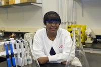 Young black woman, wearing white lab coat, leaning on lab table in lab room environment.