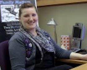 Young woman, brown hair pulled back, wearing grey sweater and purple multi-patterned scarf, sitting in front of keyboard and computer display.