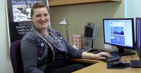 Young woman, brown hair pulled back, wearing grey sweater and purple multi-patterned scarf, sitting in front of keyboard and computer display.