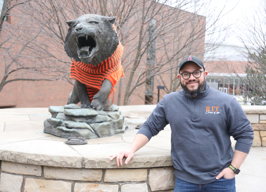 Photo of Arturo Peralta Silva, young man wearing RIT shirt and baseball cap, hand in pocket, posing by the RIT tiger statue wearing an orange stripped sweater.