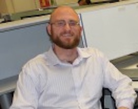 Young man, bald, dark short beard and mustache, wearing white shirt, sitting at office desk.