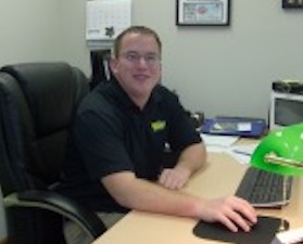 Young man, short dark hair, dark shirt, sitting in office at desk, arm extended with hand on computer mouse.