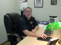 Young man, short dark hair, dark shirt, sitting in office at desk, arm extended with hand on computer mouse.