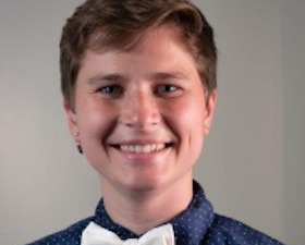 Young woman, portrait style, short brown hair, dark blue shirt and white bow tie.