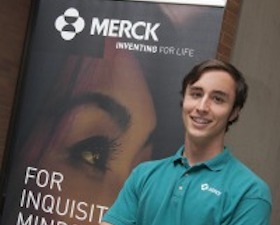 Young man, short dark hair, green shirt, standing in front of sign for Merck company.