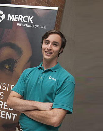 Young man, short dark hair, green shirt, standing in front of sign for Merck company.