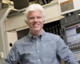 Young man, very light blond short hair, plaid shirt, standing in room of technical monitoring equipment.