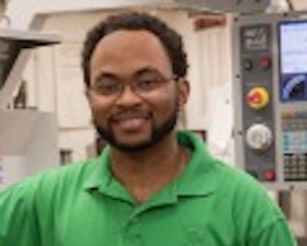 Young man, dark skin, glasses, short dark mustache and beard, wearing light green polo shirt, standing in front of technical monitoring equipment.