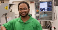 Young man, dark skin, glasses, short dark mustache and beard, wearing light green polo shirt, standing in front of technical monitoring equipment.