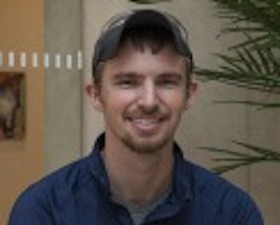 Young man, dark hair, short mustache and beard, standing near plant.