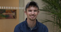 Young man, dark hair, short mustache and beard, standing near plant.