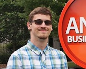 Young man, dark hair, sunglasses, wearing plaid shirt, standing by sign that says ANNEX Business Media.