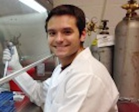 Young man, short dark hair, wearing lab coat, in lab and holding pipette.