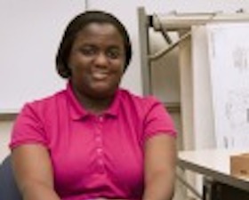 Young woman, dark skin, pink top, sitting at computer with blueprints on display monitor.