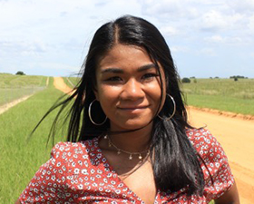 Young woman, long dark hair, hoop earrings, red top with small white flowers, standing outside in field.