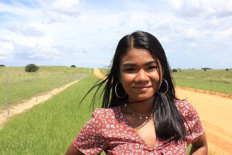 Young woman, long dark hair, hoop earrings, red top with small white flowers, standing outside in field.