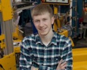 Young man, blond hair, arms crossed, plaid shirt, in front of engineering equipment.