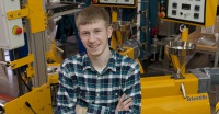 Young man, blond hair, arms crossed, plaid shirt, in front of engineering equipment.