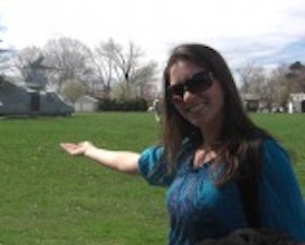 Young woman, long brown hair, sunglasses, bluish-purple blouse, standing outside and holding out arm to indicate landed military helicopter behind her.