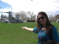 Young woman, long brown hair, sunglasses, bluish-purple blouse, standing outside and holding out arm to indicate landed military helicopter behind her.