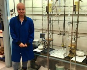 Young woman, tan head covering, glasses, wearing dark blue lab coat, standing next to lab equipment.