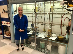 Young woman, tan head covering, glasses, wearing dark blue lab coat, standing next to lab equipment.