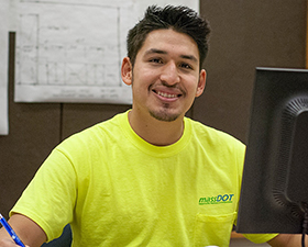 Young man, short dark hair, yellow shirt, sitting in front of computer monitor.