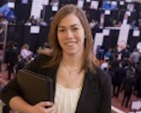 Young woman, long brown hair, white top and dark suit jacket, holding notebook, standing at top of stairs with trade show and people below.