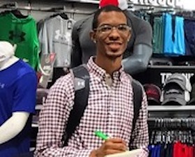 Young man, dark skin and glasses, standing amid sports apparel.