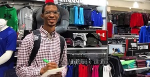 Young man, dark skin and glasses, standing amid sports apparel.