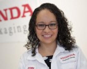 Young woman, dark curly hair, wearing lab coat, standing in front of sign that says: HONDA Packaging.
