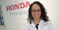 Young woman, dark curly hair, wearing lab coat, standing in front of sign that says: HONDA Packaging.